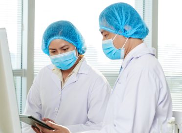 Group of hard-working researchers wearing medical masks and white coats sitting at laboratory bench and studying results of conducted experiment with help of digital tablet